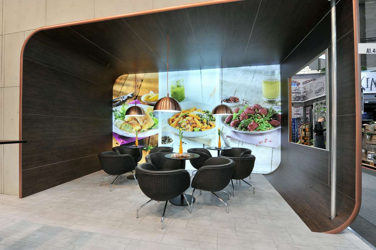 Modernes Messedesign mit Holzelementen, Sitzgruppen mit schwarzen Ledersesseln und der Abbildung von Essen im Hintergrund.